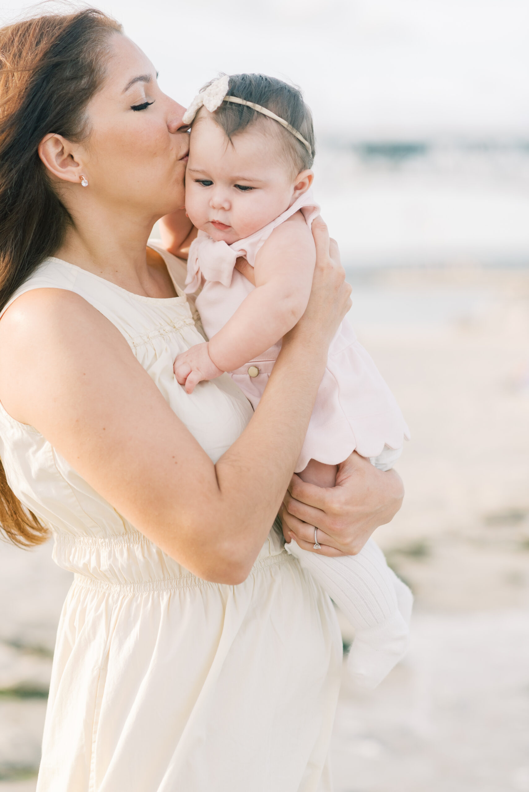 mom wearing a white dress holding and kissing baby on the bead in the water at Virginia Beach